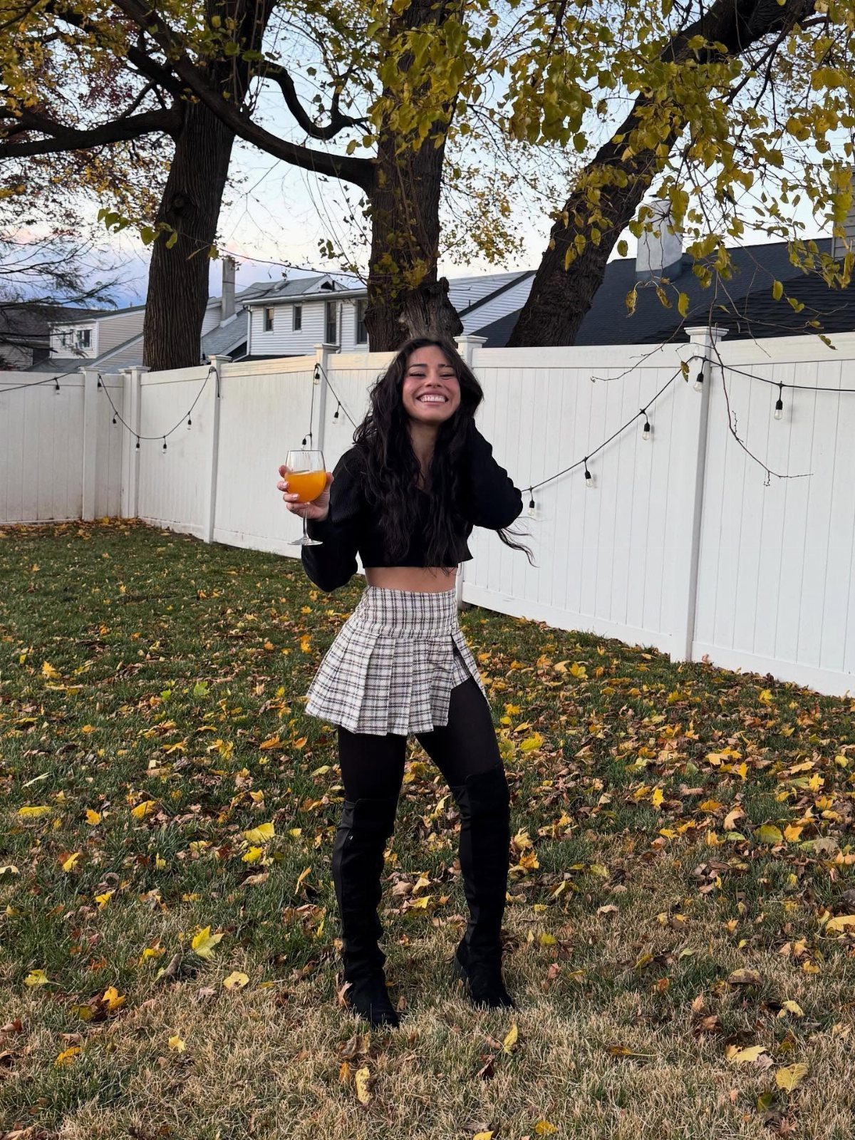 Smiling woman in a plaid skirt, black crop top, and thigh-high boots standing in a yard.