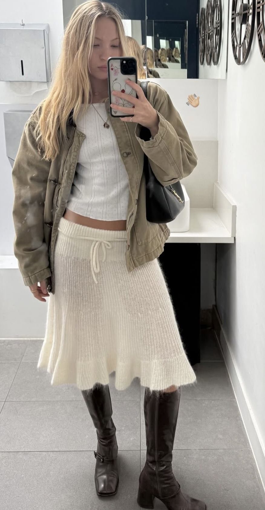 Blonde woman in a cream knit skirt, olive jacket, and tall brown boots taking a mirror selfie.