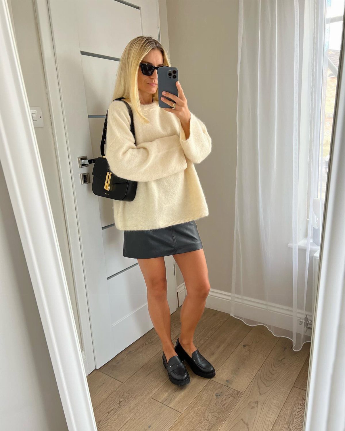 A woman wearing a cream knit sweater and a black leather mini skirt with black loafers.