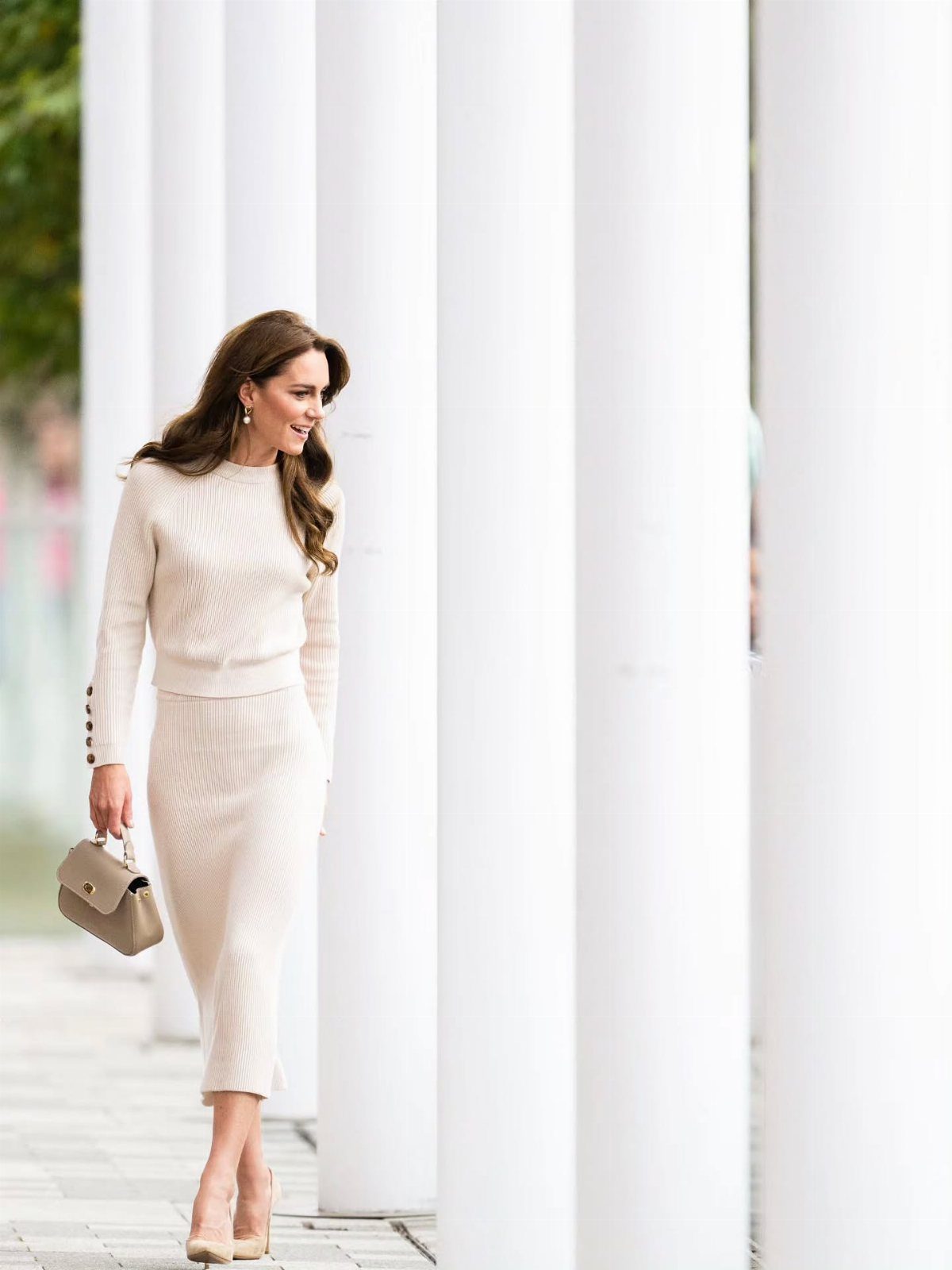 Woman in a cream ribbed knit midi skirt and matching top walking past white pillars.