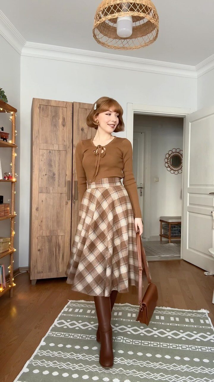 Woman wearing a brown tie-neck top, plaid midi skirt, and tall brown boots standing in a room.