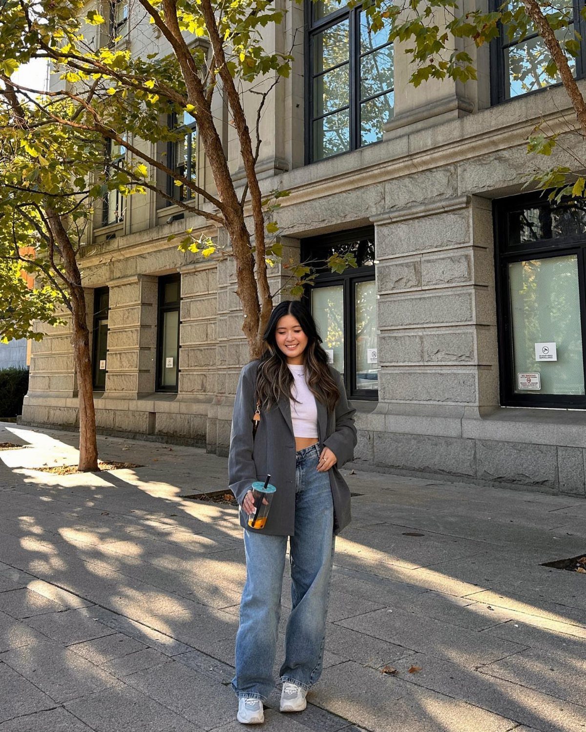 A smiling woman wearing a grey blazer, white crop top, and wide-leg blue jeans standing outdoors.