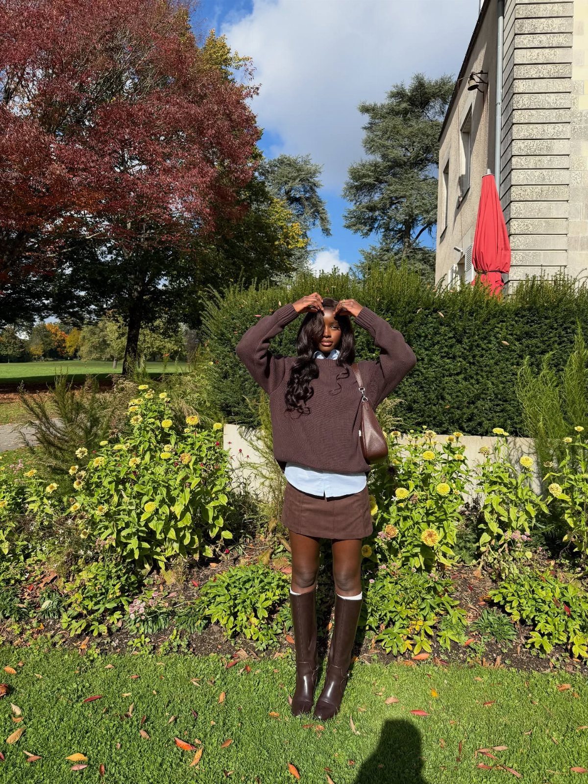A young woman posing in a brown sweater, skirt, and tall boots amidst autumn garden greenery.