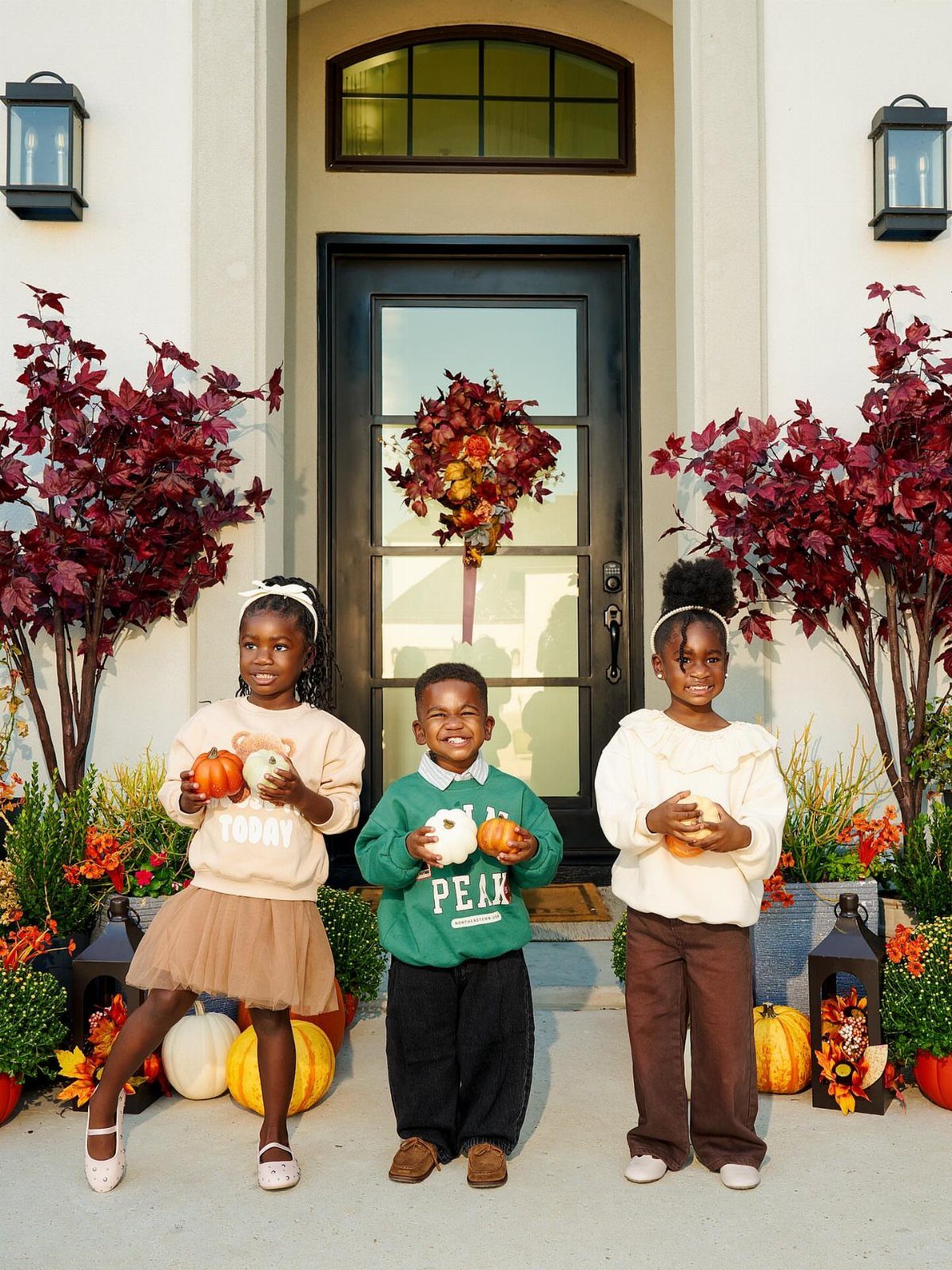 Three children in fall clothing pose with pumpkins in front of a decorated doorway.