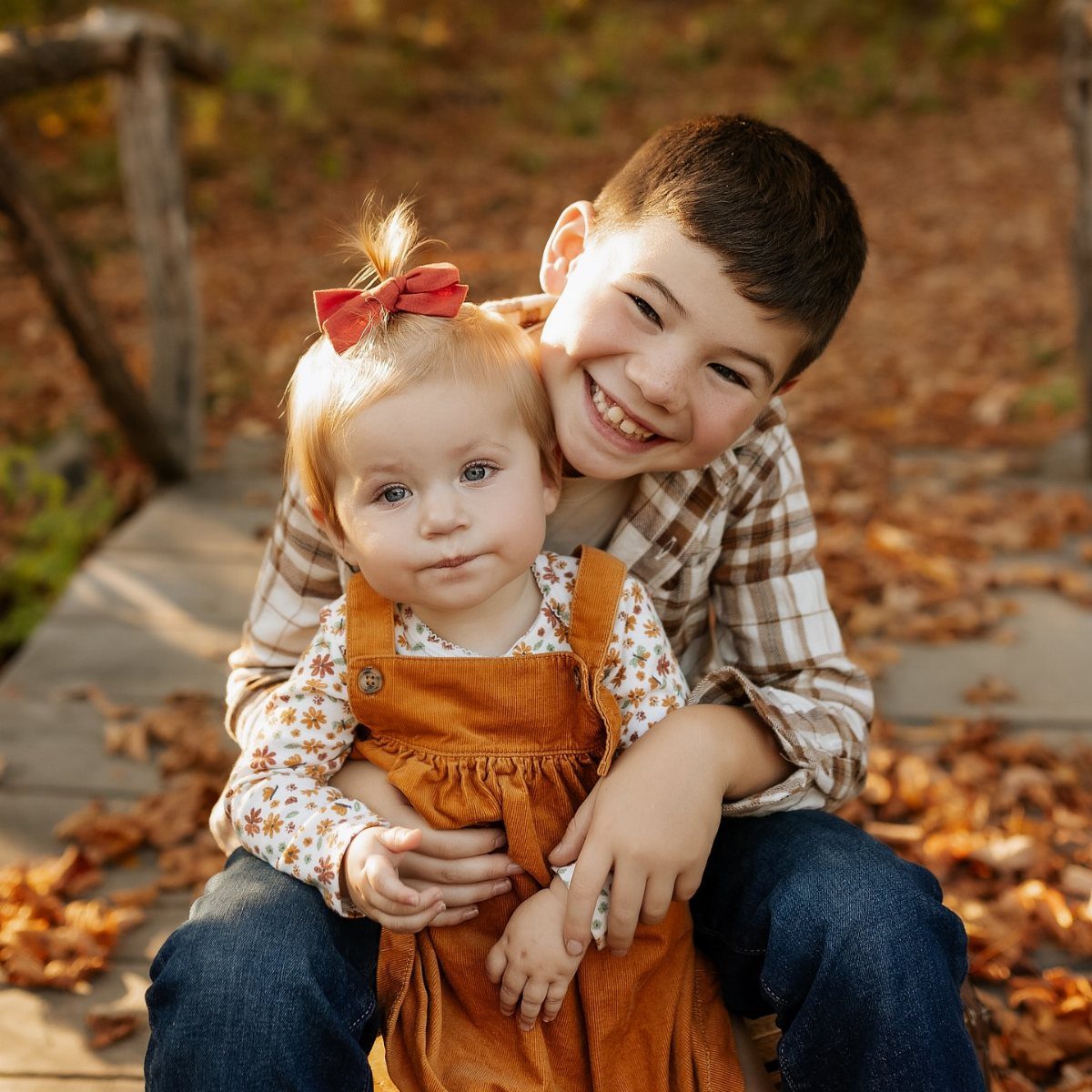 A smiling boy hugs a toddler girl wearing a red hair bow and orange corduroy overalls.