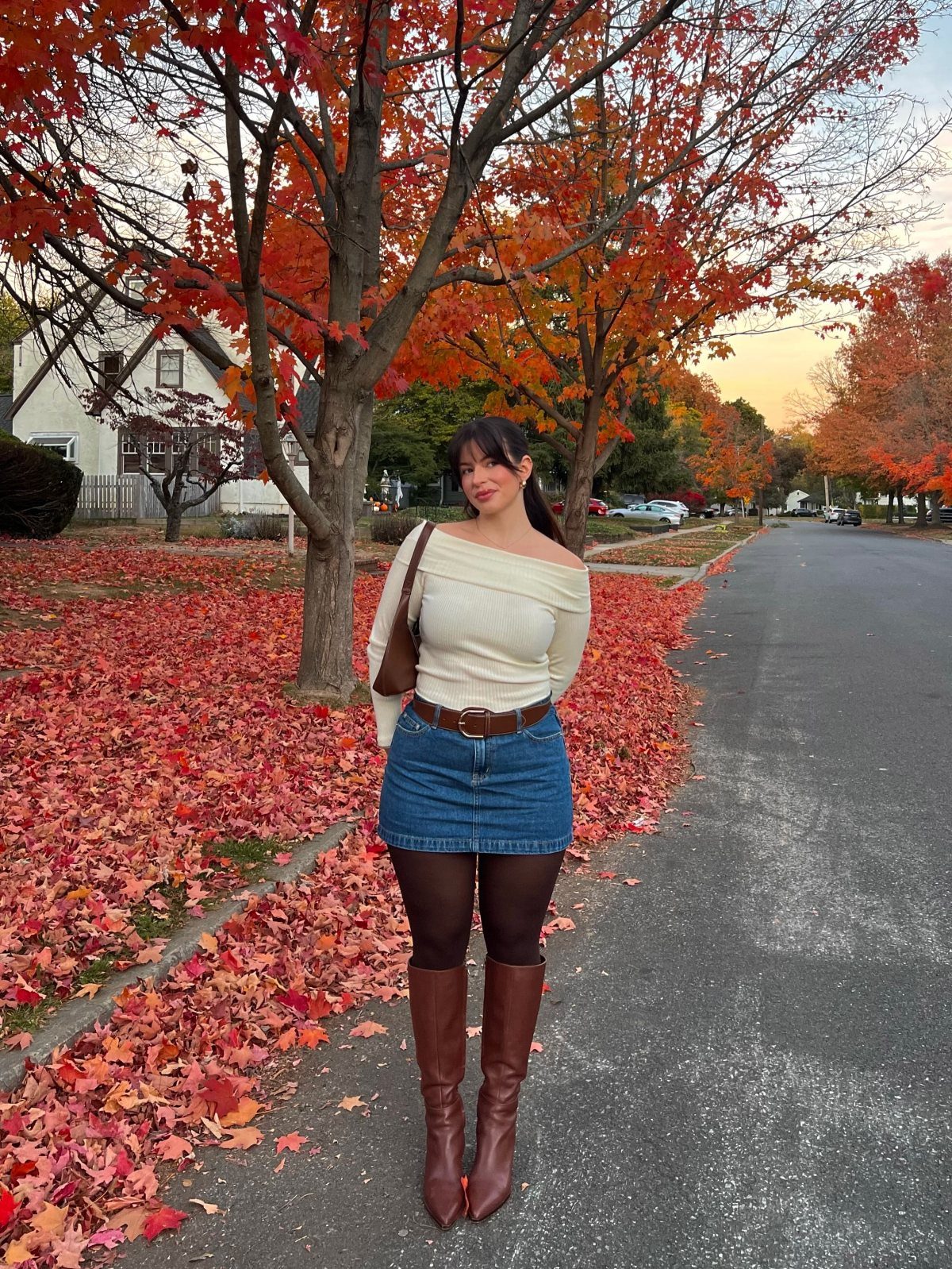 A woman wearing a cream top, denim skirt, and brown boots stands among autumn leaves.