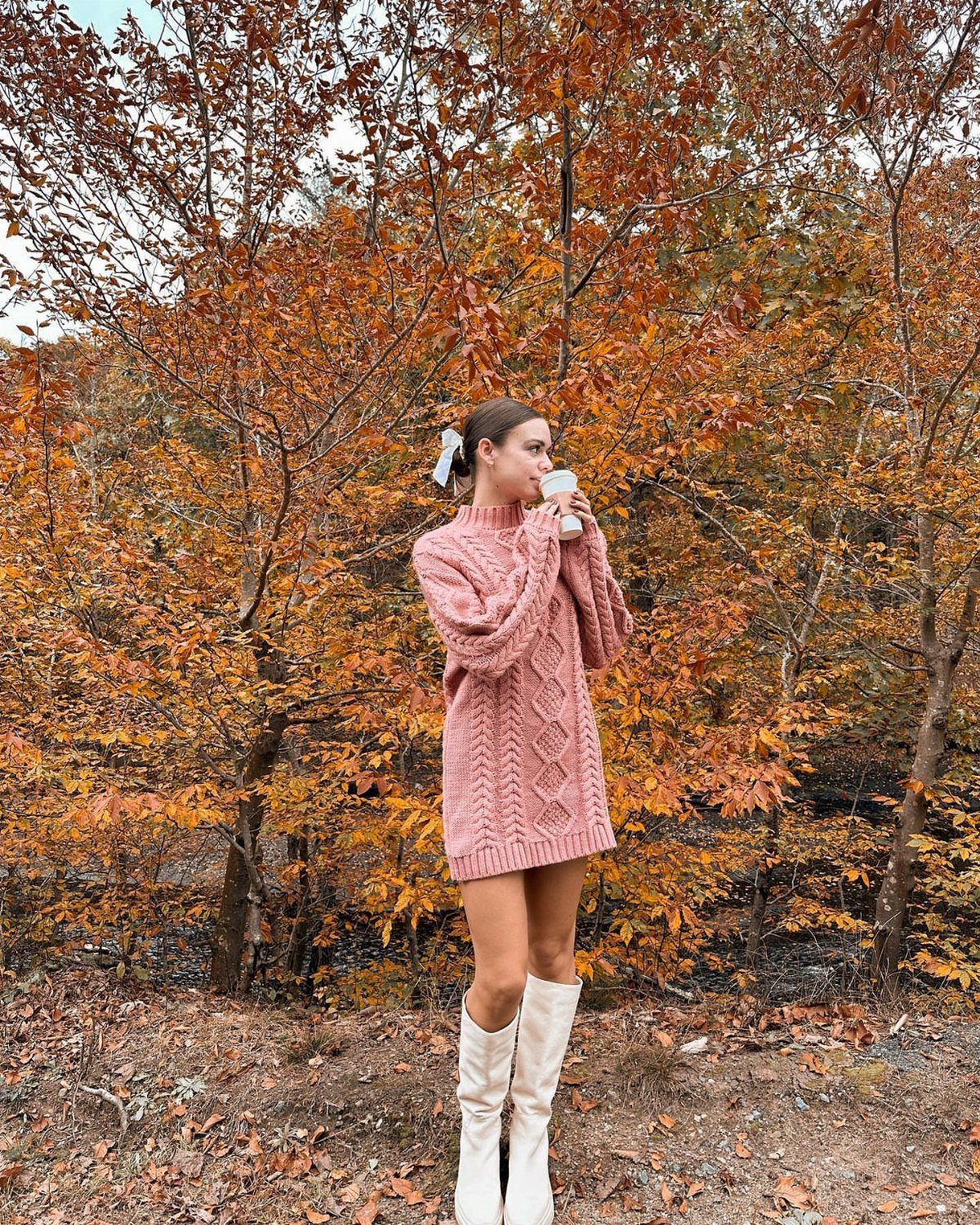 A woman in a pink cable-knit sweater dress and white boots standing among autumn trees.