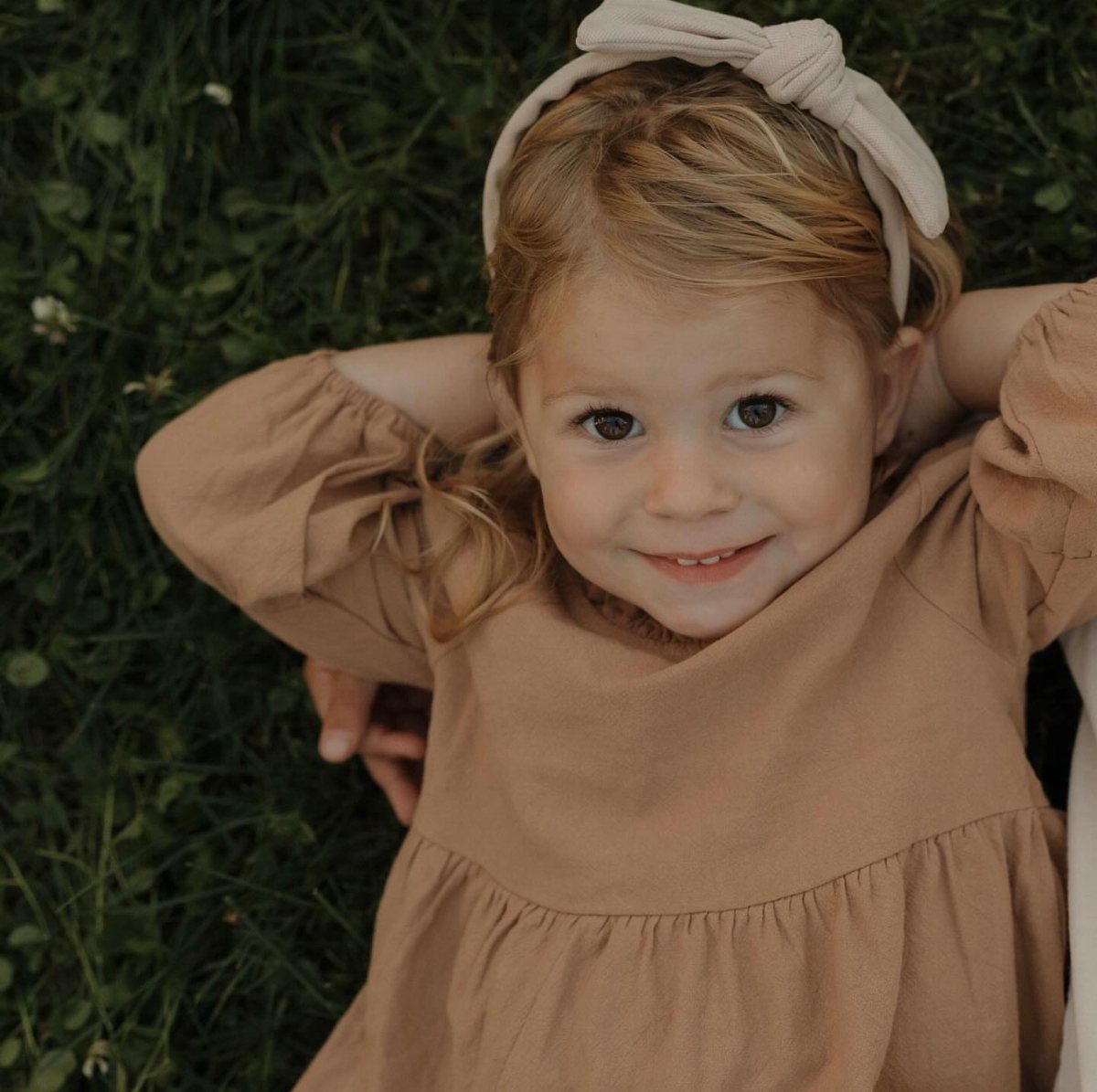 A smiling young girl in a tan dress lying on green grass wearing a headband.