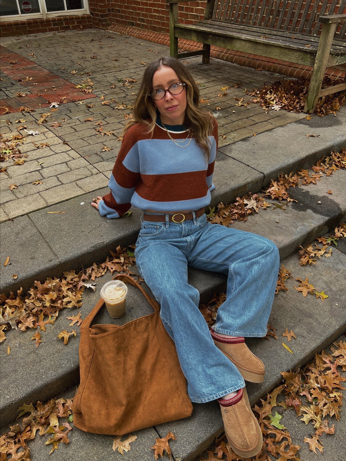A woman wearing a striped sweater and blue jeans sits on stone steps with autumn leaves.