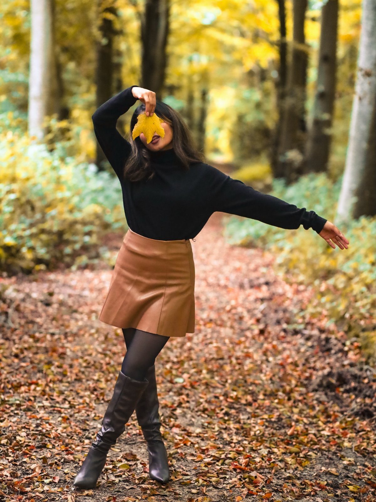 Woman in a black sweater and tan leather skirt posing in an autumn forest.