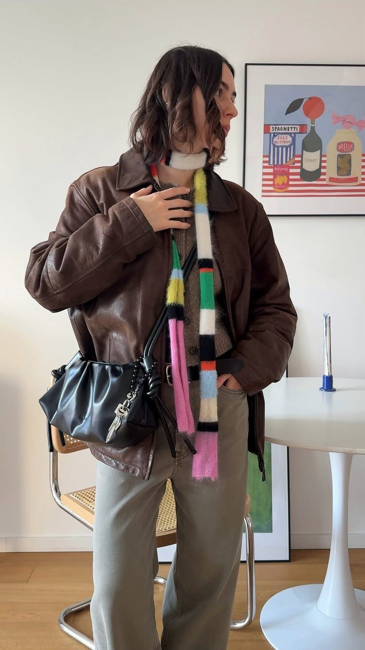 Woman wearing a brown leather jacket, tan top, olive trousers, and a colorful striped scarf.