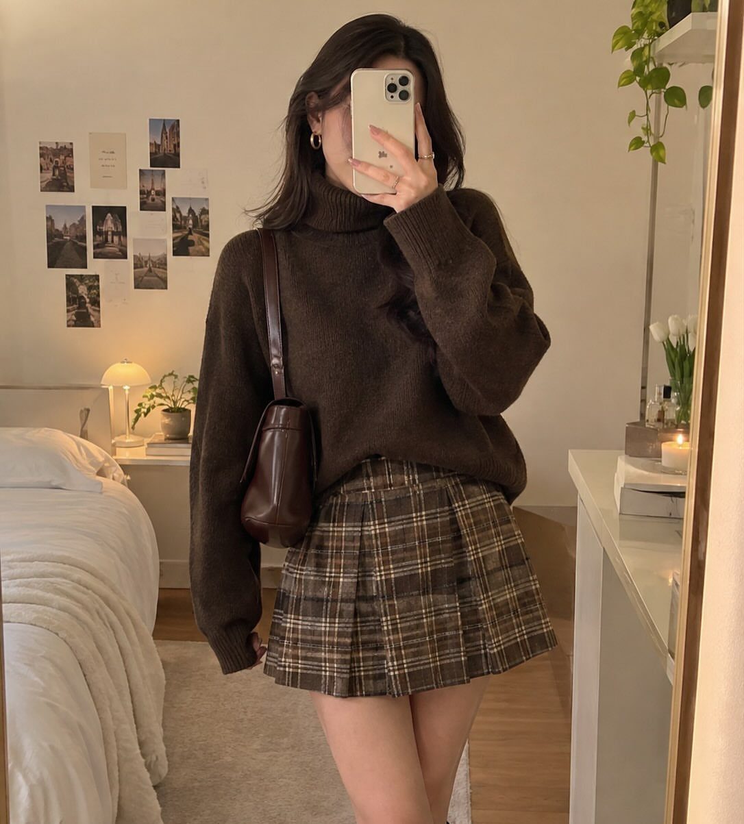 A person wearing a dark brown turtleneck sweater and a plaid pleated skirt standing in a bedroom.