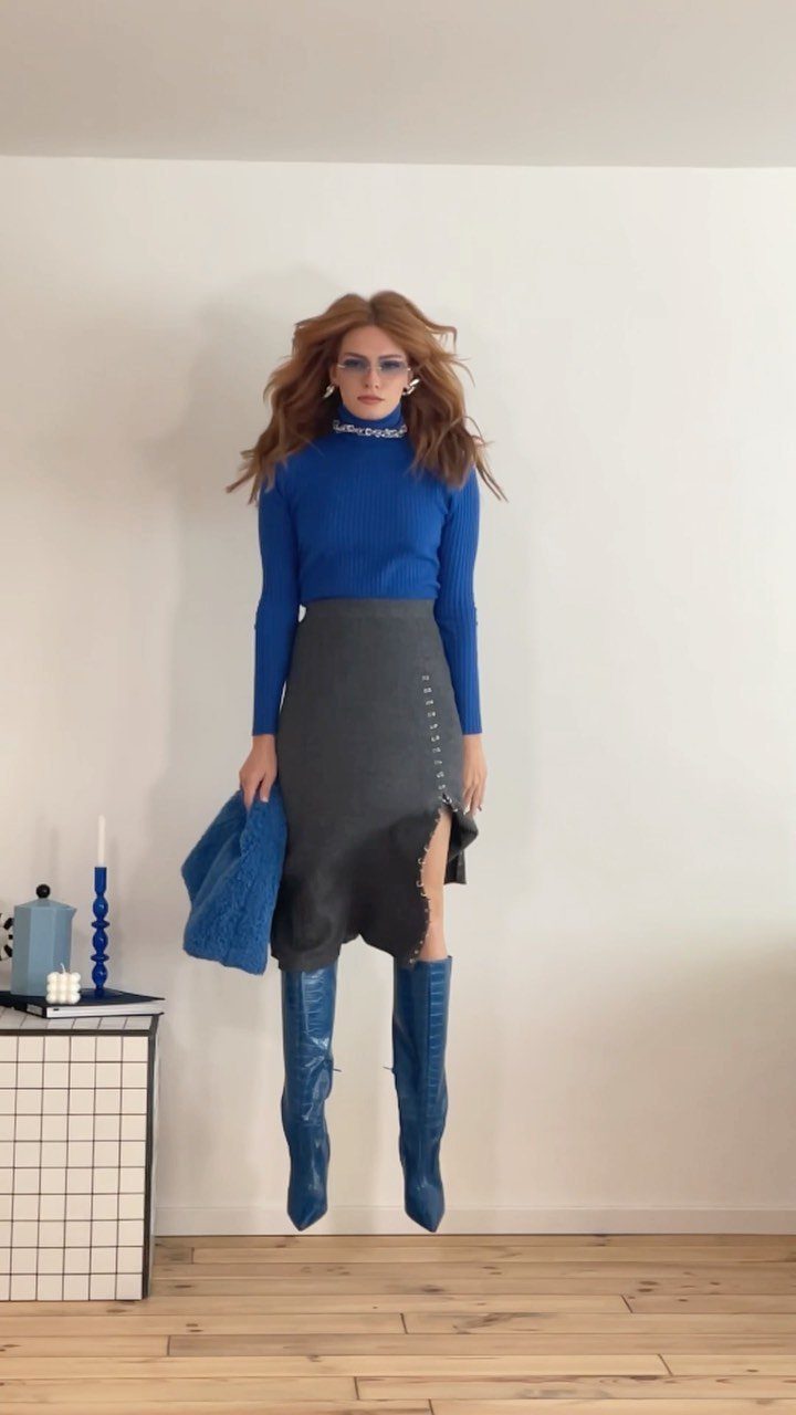 A woman in a blue turtleneck, grey skirt, and blue boots posing against a white wall.