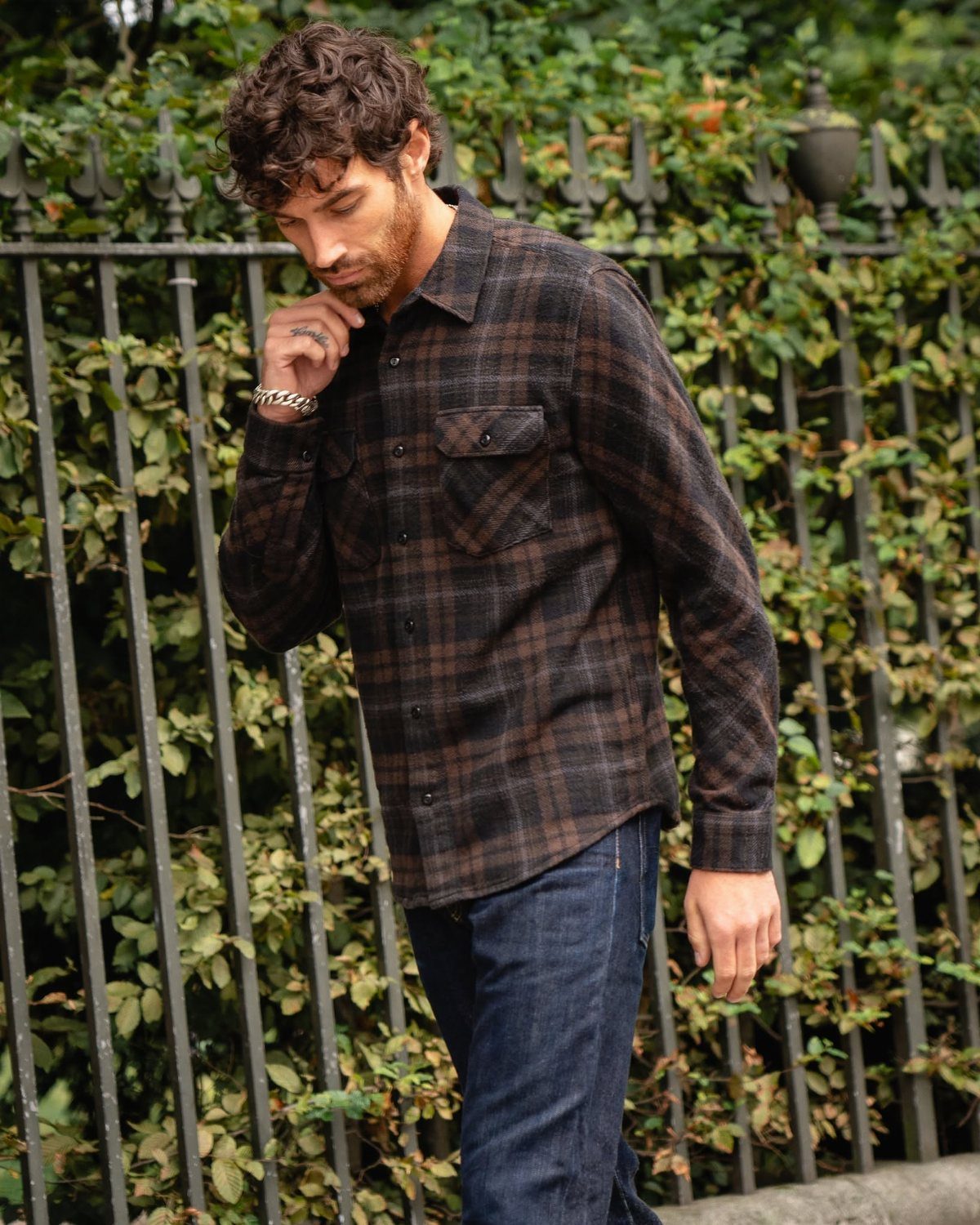 A man in a plaid button-down shirt and jeans walking past a garden fence.