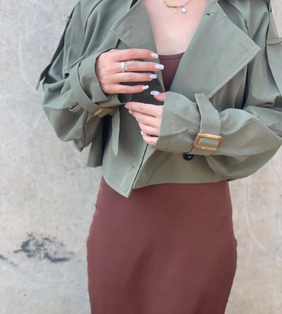 A person wearing a cropped olive green trench coat over a brown dress.