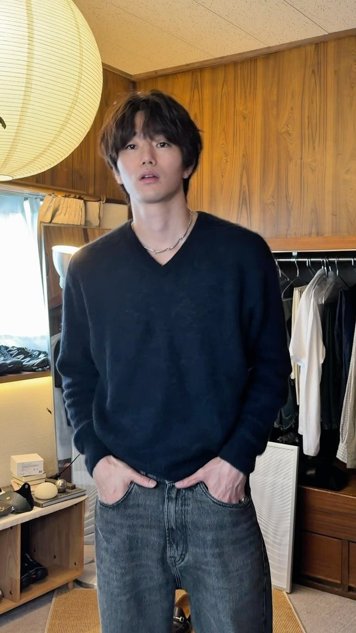 A young man posing in a dark navy V-neck knit sweater and grey jeans.