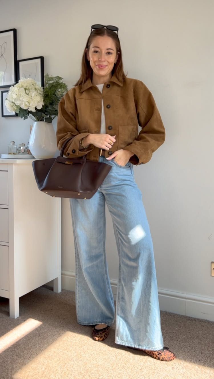 Woman posing in a brown suede jacket, white top, and wide-leg blue jeans.