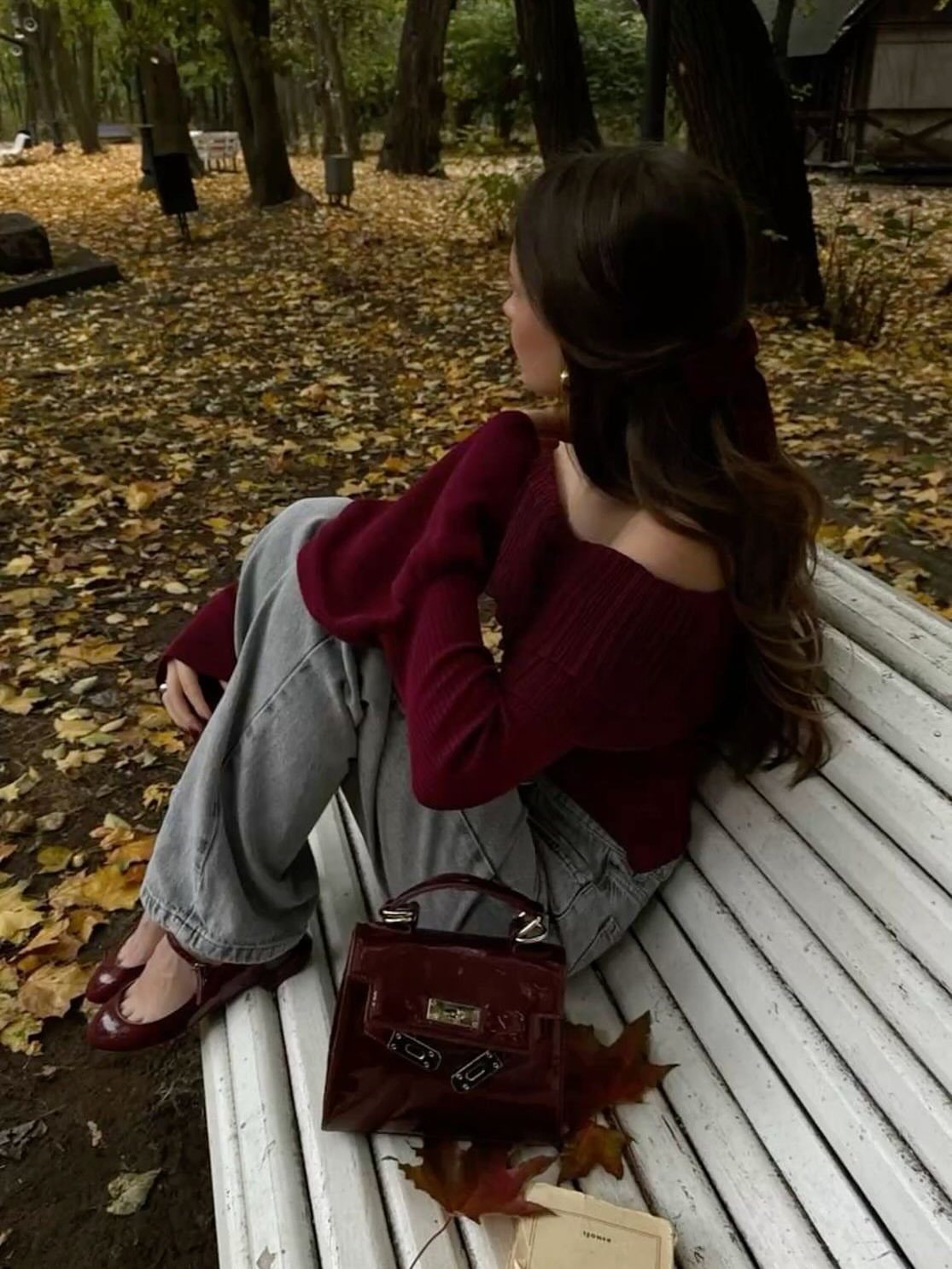 Woman in burgundy sweater and jeans sitting on a white bench amidst autumn leaves.