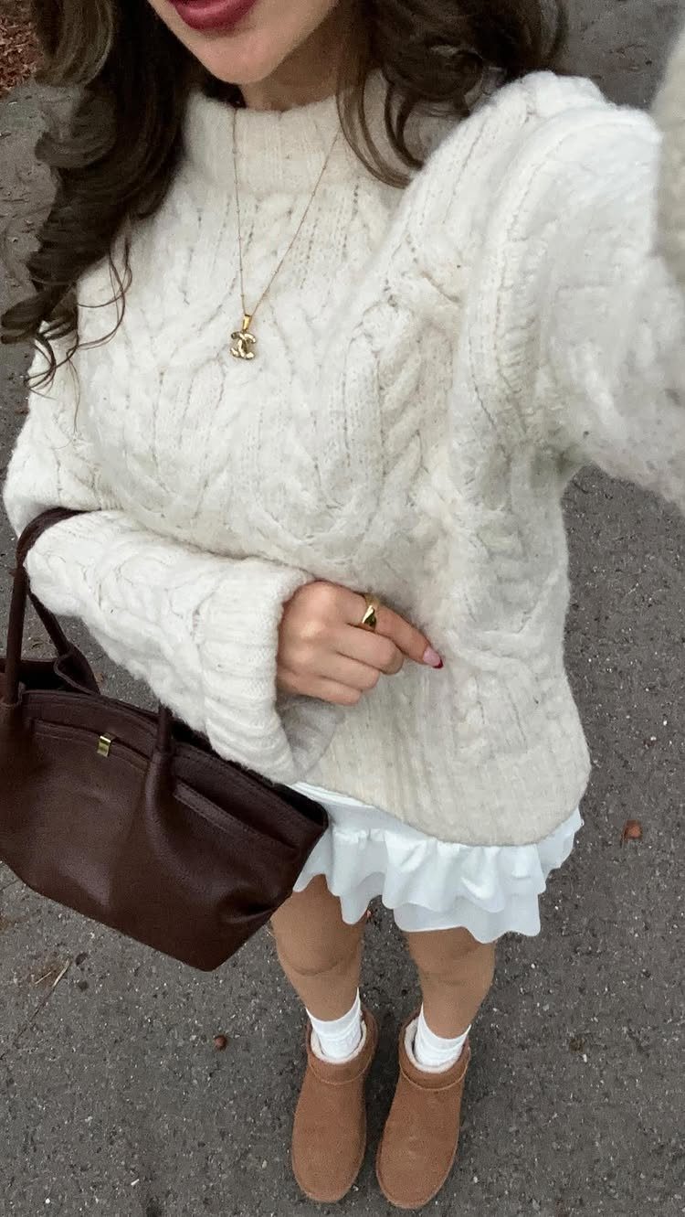 A woman wearing a white cable-knit sweater, ruffled skirt, and tan clogs holding a brown handbag.