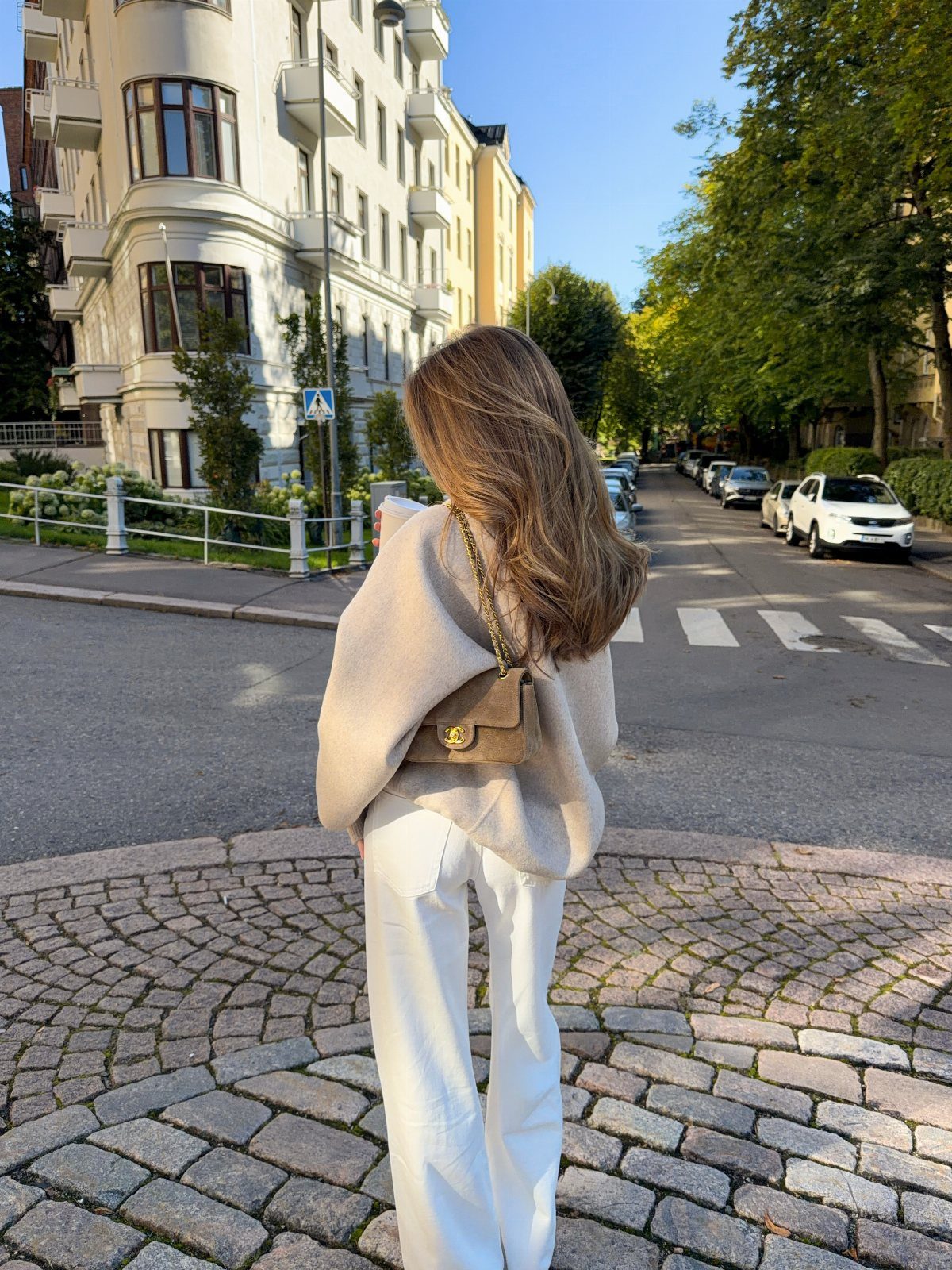 Woman in cream sweater and white pants carrying a tan leather crossbody bag on a cobblestone street.