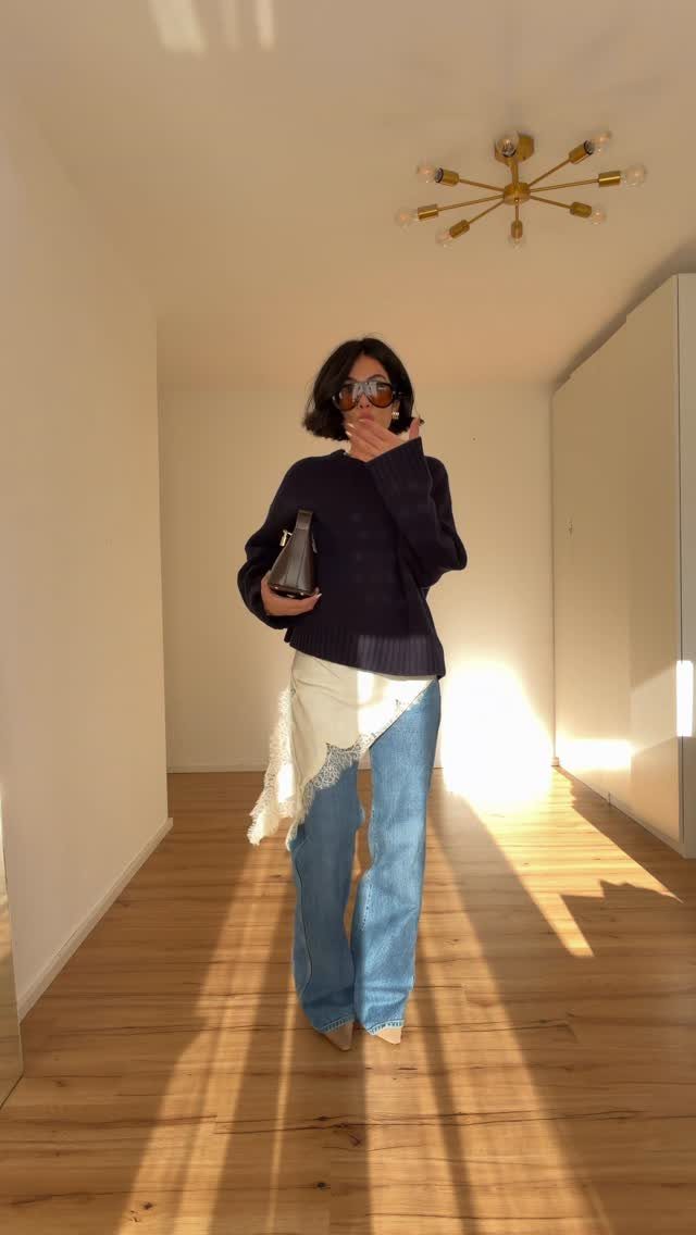 Woman in a navy sweater and blue jeans standing in a sunlit hallway.