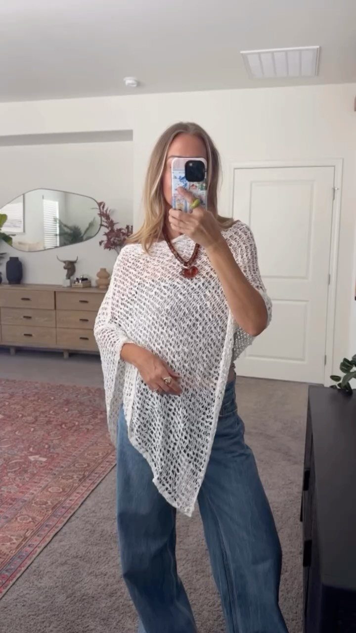 Woman in a white crochet poncho and wide-leg blue denim jeans taking a mirror selfie.