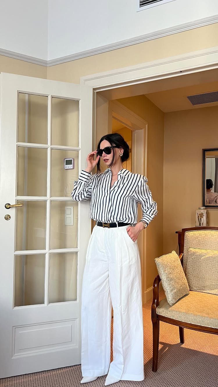 Woman in a striped shirt and white wide-leg trousers standing in a doorway.