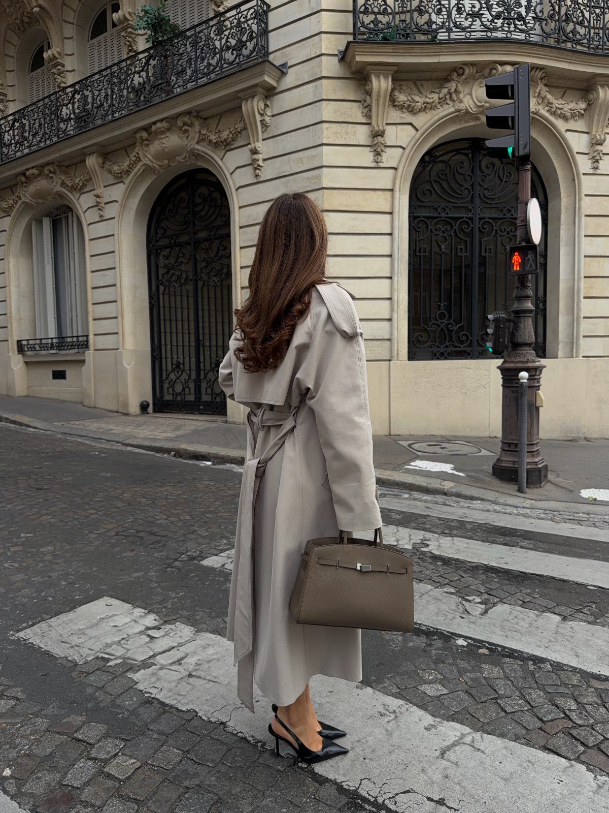 A woman in a long beige trench coat and black slingbacks walking on a city street.
