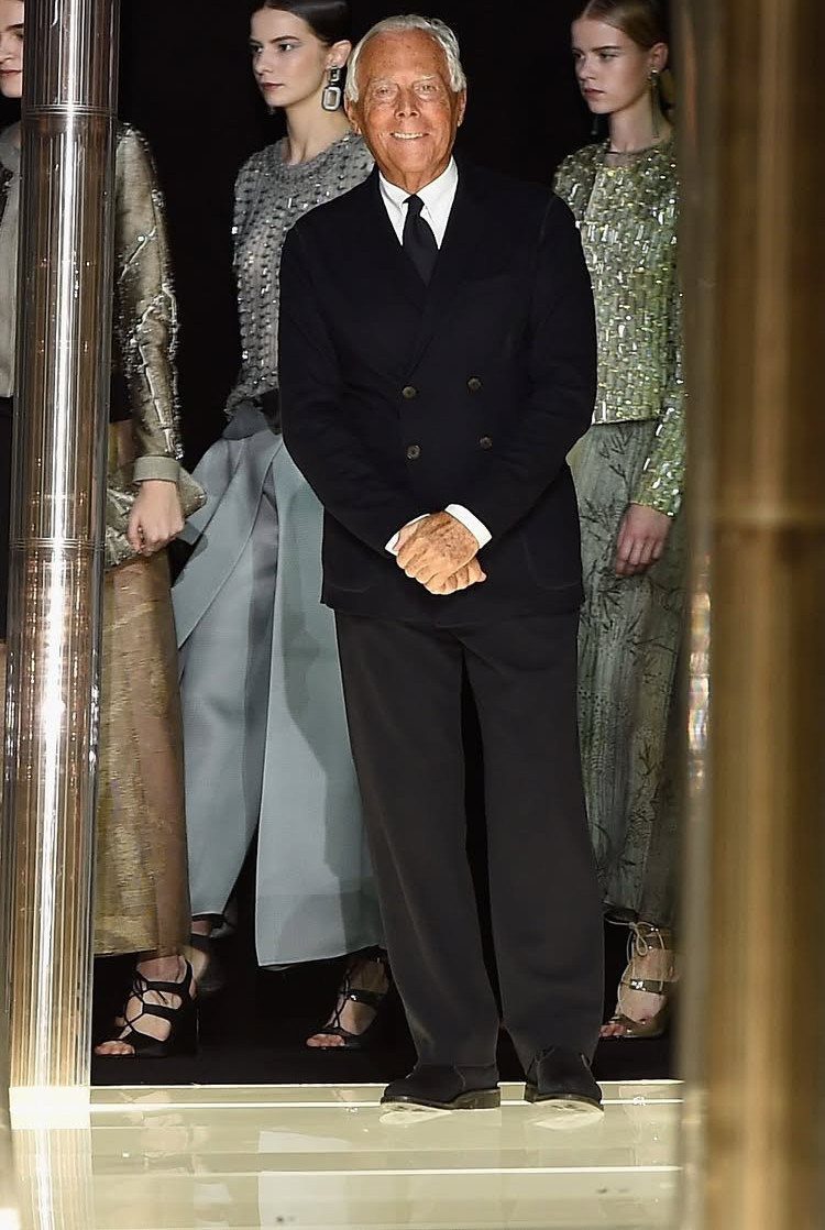 Giorgio Armani smiling in a dark navy suit while standing before fashion models.