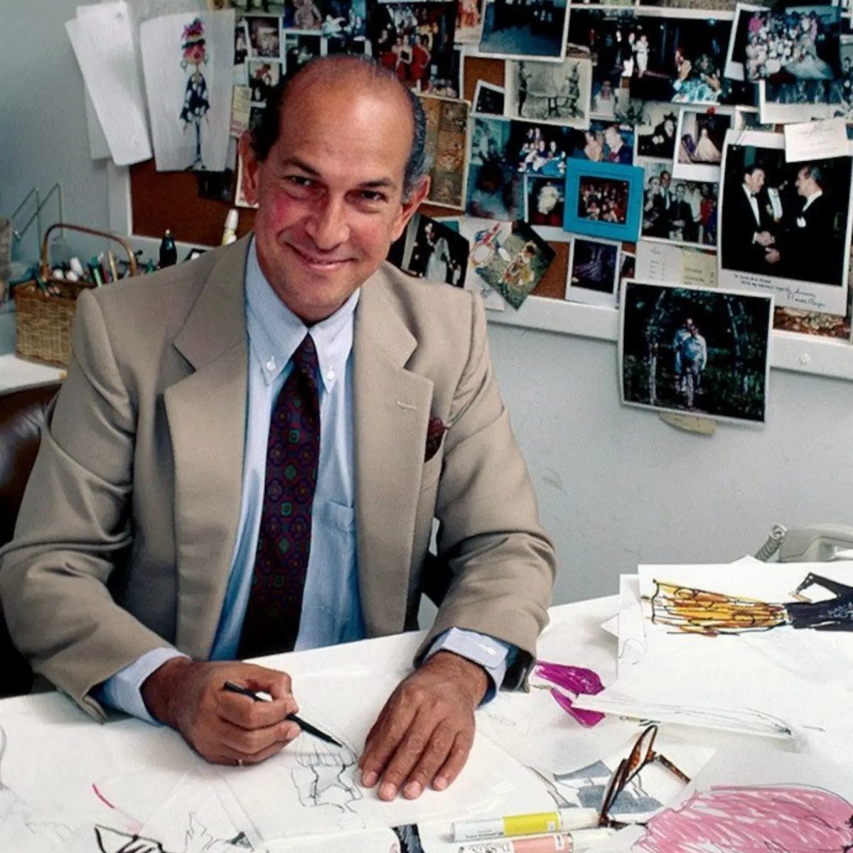 Oscar de la Renta sitting at a desk sketching fashion designs with photographs in the background.