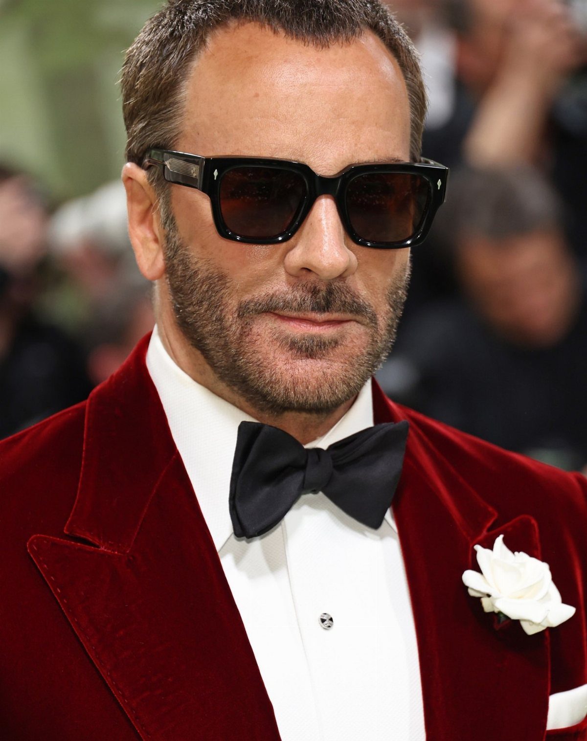 Tom Ford wearing black sunglasses and a red velvet tuxedo jacket with a white boutonniere.