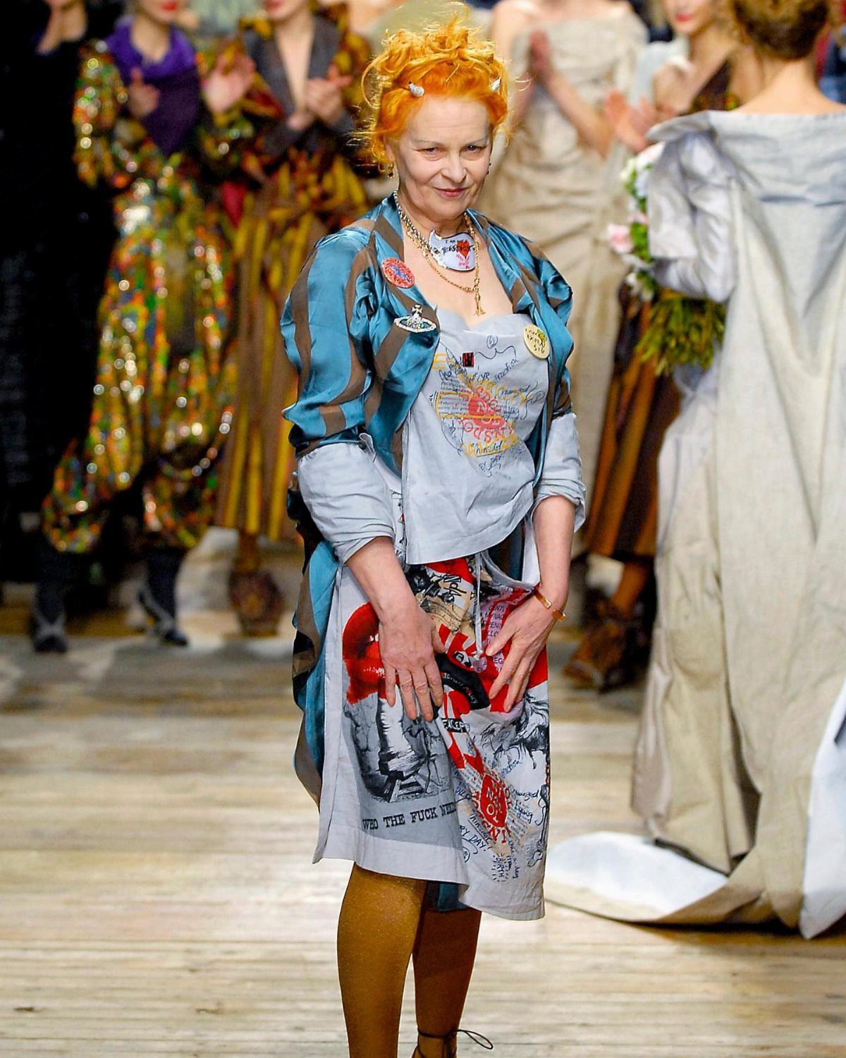 Vivienne Westwood with orange hair walking a fashion runway wearing eclectic layered clothing.
