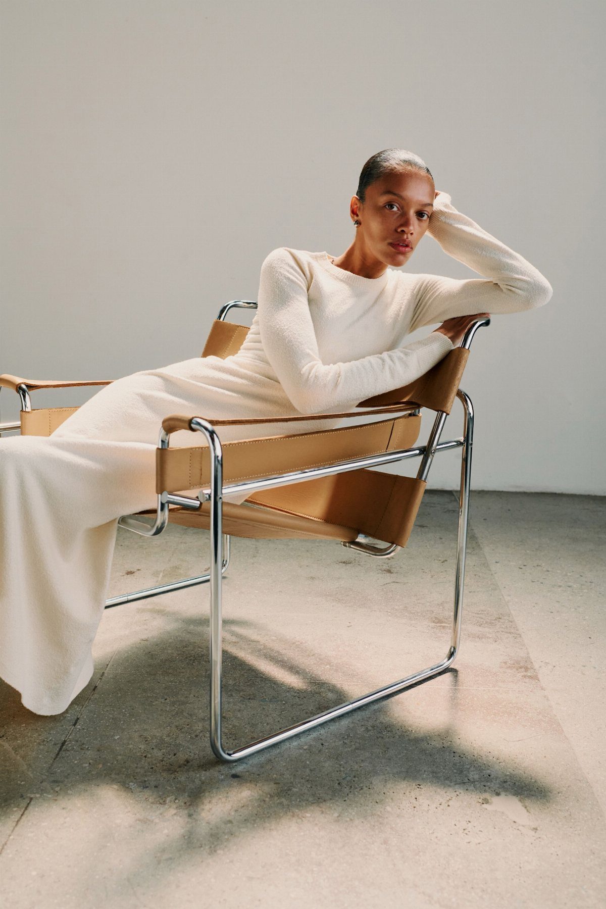 Woman in a white long-sleeved dress sitting in a leather chair against a white wall.