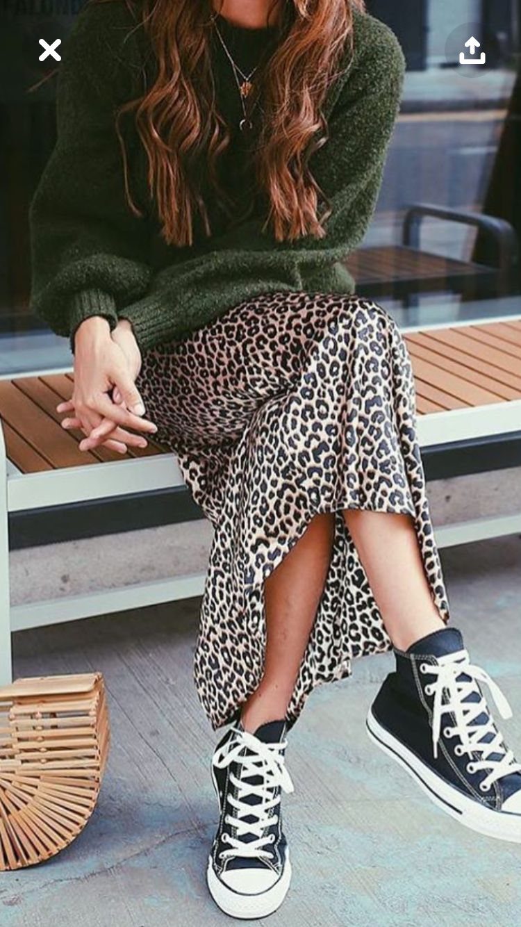 A person wearing a green sweater, leopard print long skirt, and black high-top sneakers sitting on a bench.