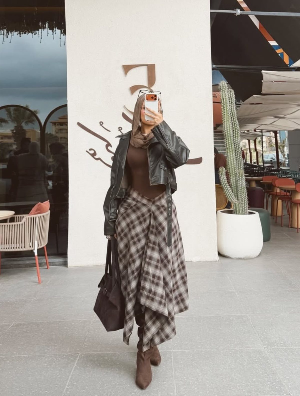 A person posing in a leather jacket, brown bodysuit, and long plaid skirt.