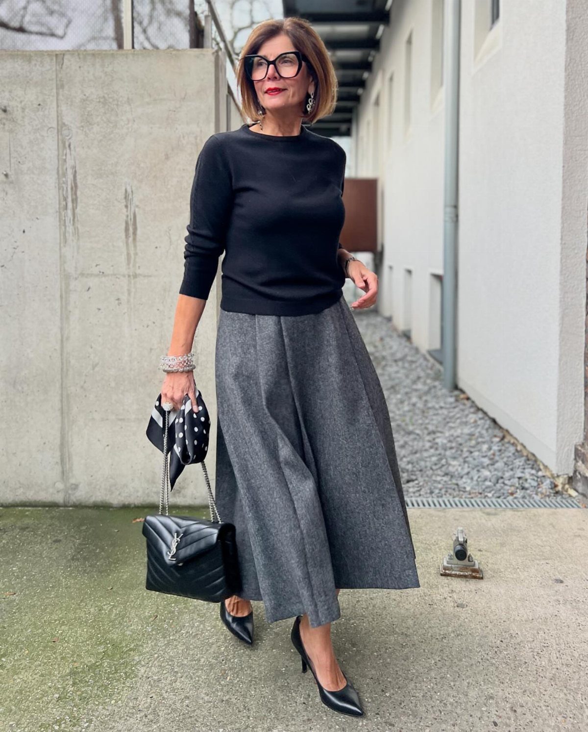 A mature woman wearing a black top and grey long skirt posing outdoors.