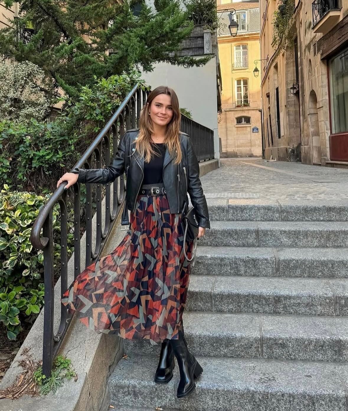 A woman wearing a leather jacket and patterned long skirt standing on stone stairs.