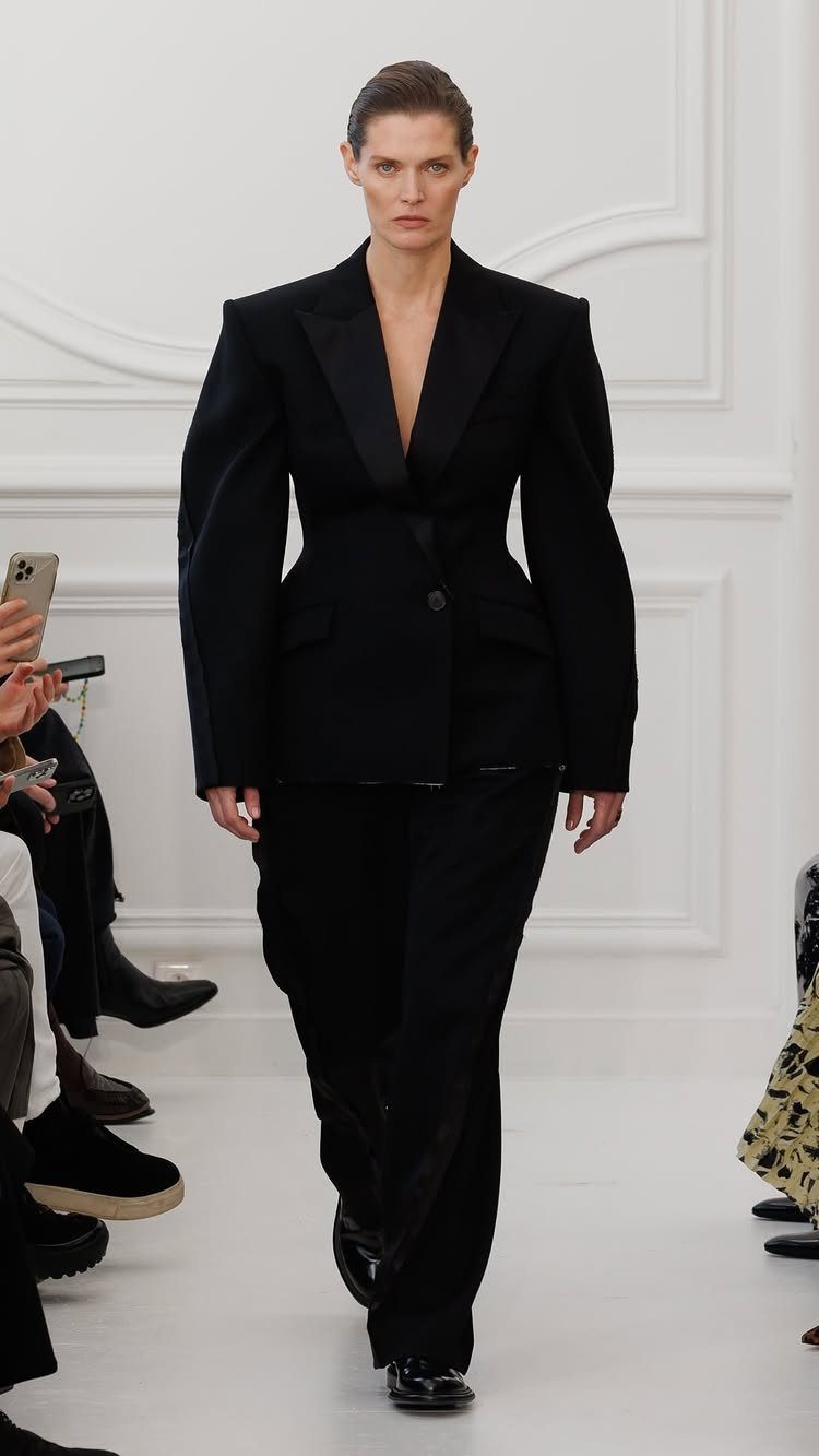 Model walking a runway in a black cinched blazer and wide-leg trousers.
