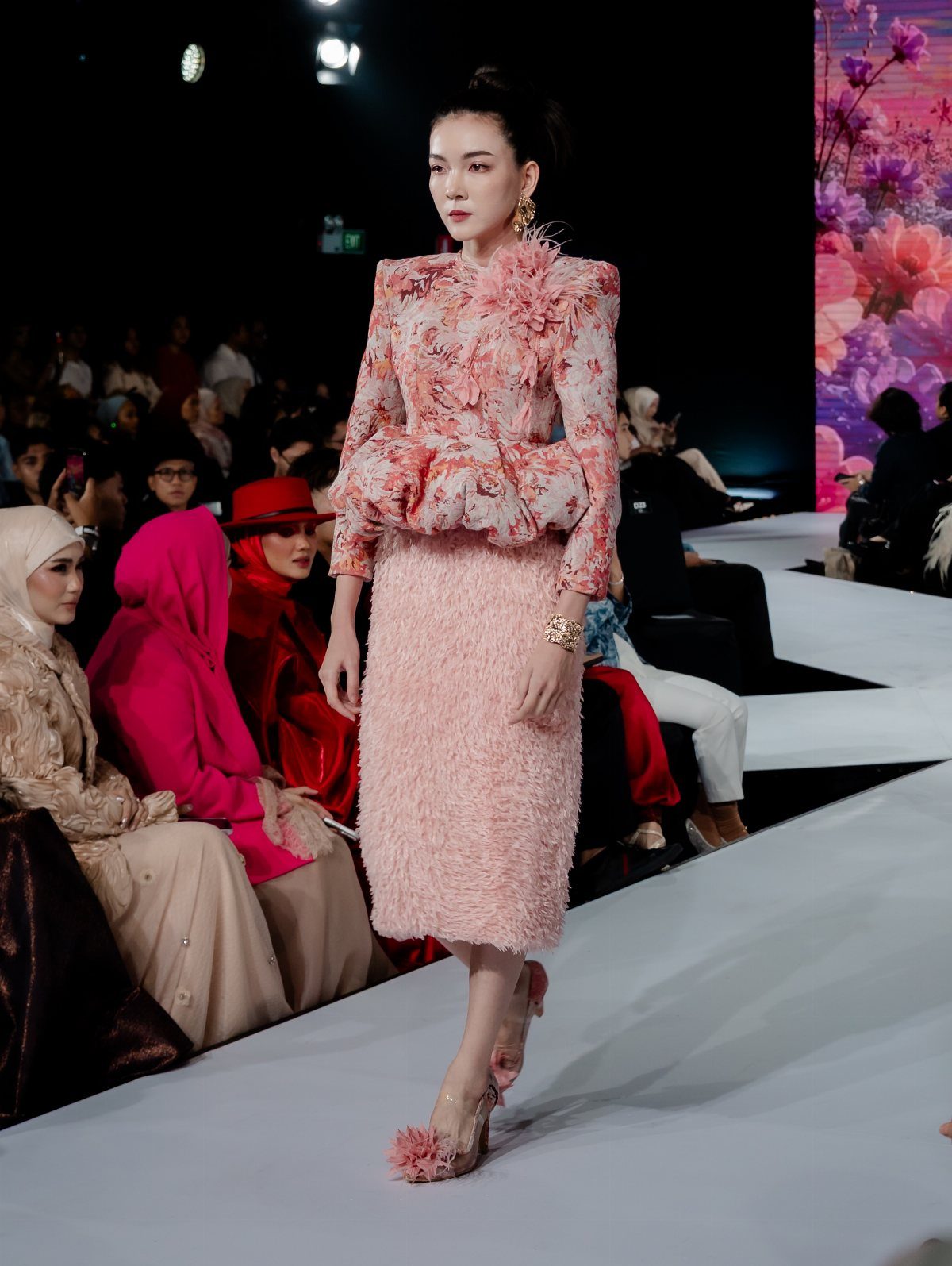 Model walking a runway in a pink floral blouse and textured pink skirt.