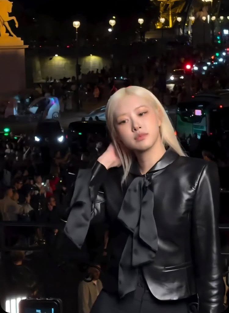 Blonde woman posing in a black leather jacket with a large satin bow at night.