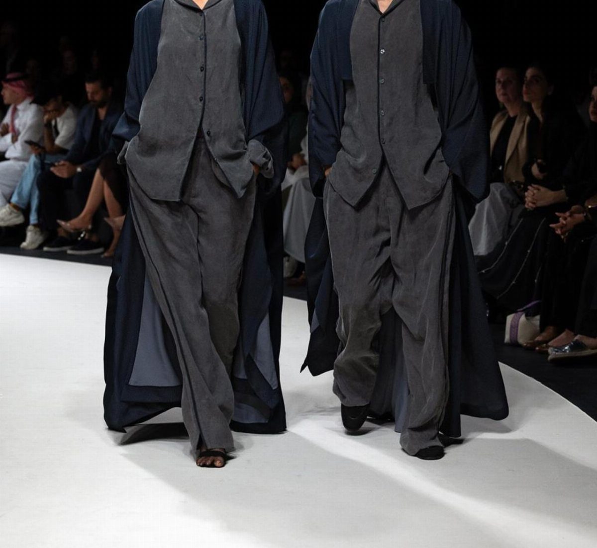 Two models walking a runway wearing matching grey waistcoats, trousers, and long navy blue coats.