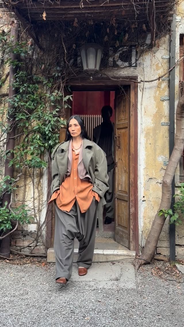 Woman wearing oversized olive coat, orange shirt, and voluminous grey trousers walking through a doorway.