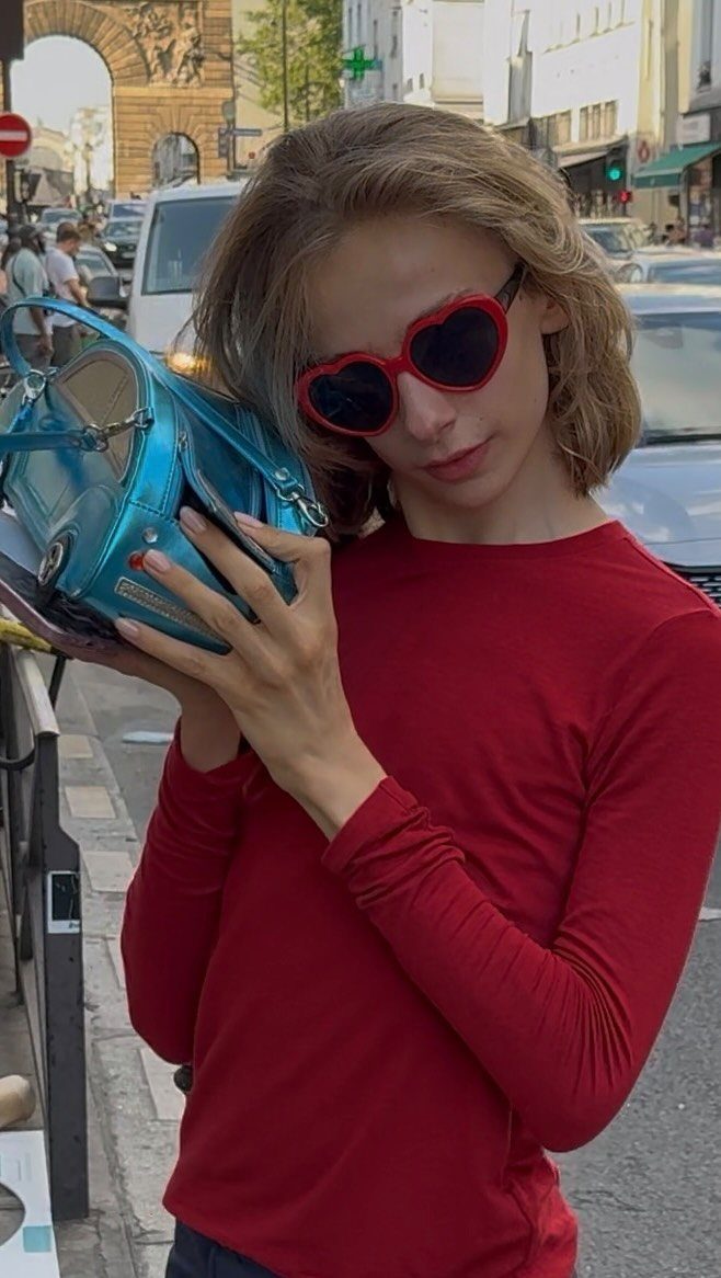 Person in red clothing and heart sunglasses holding a bright blue handbag on a city street.