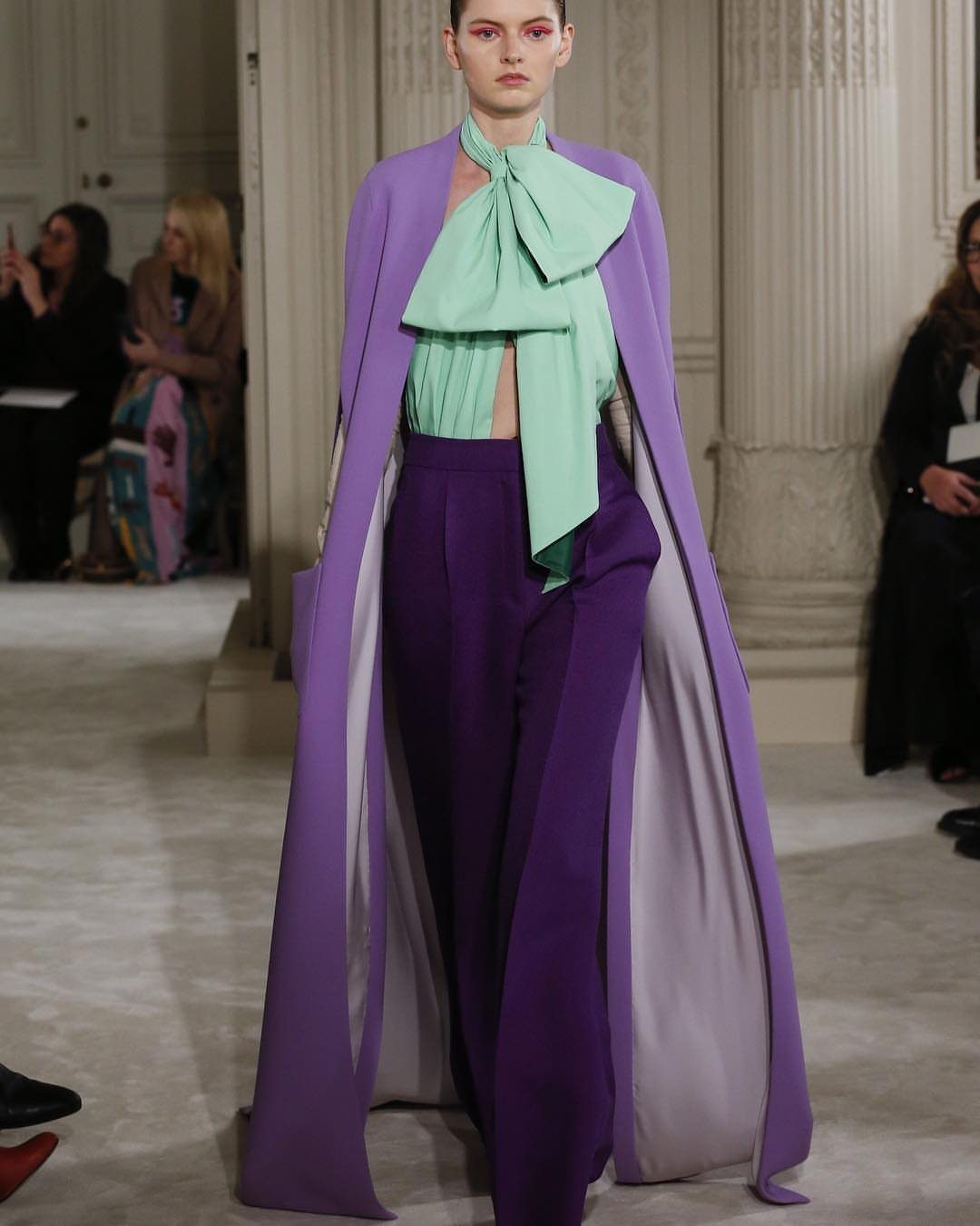 Model walking a runway in a mint green bow top, purple trousers, and a long lavender coat.