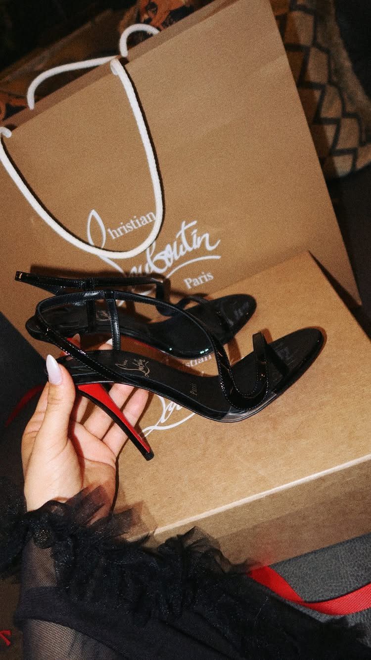 Black patent leather stiletto sandals with iconic red soles held in front of a designer shopping bag.
