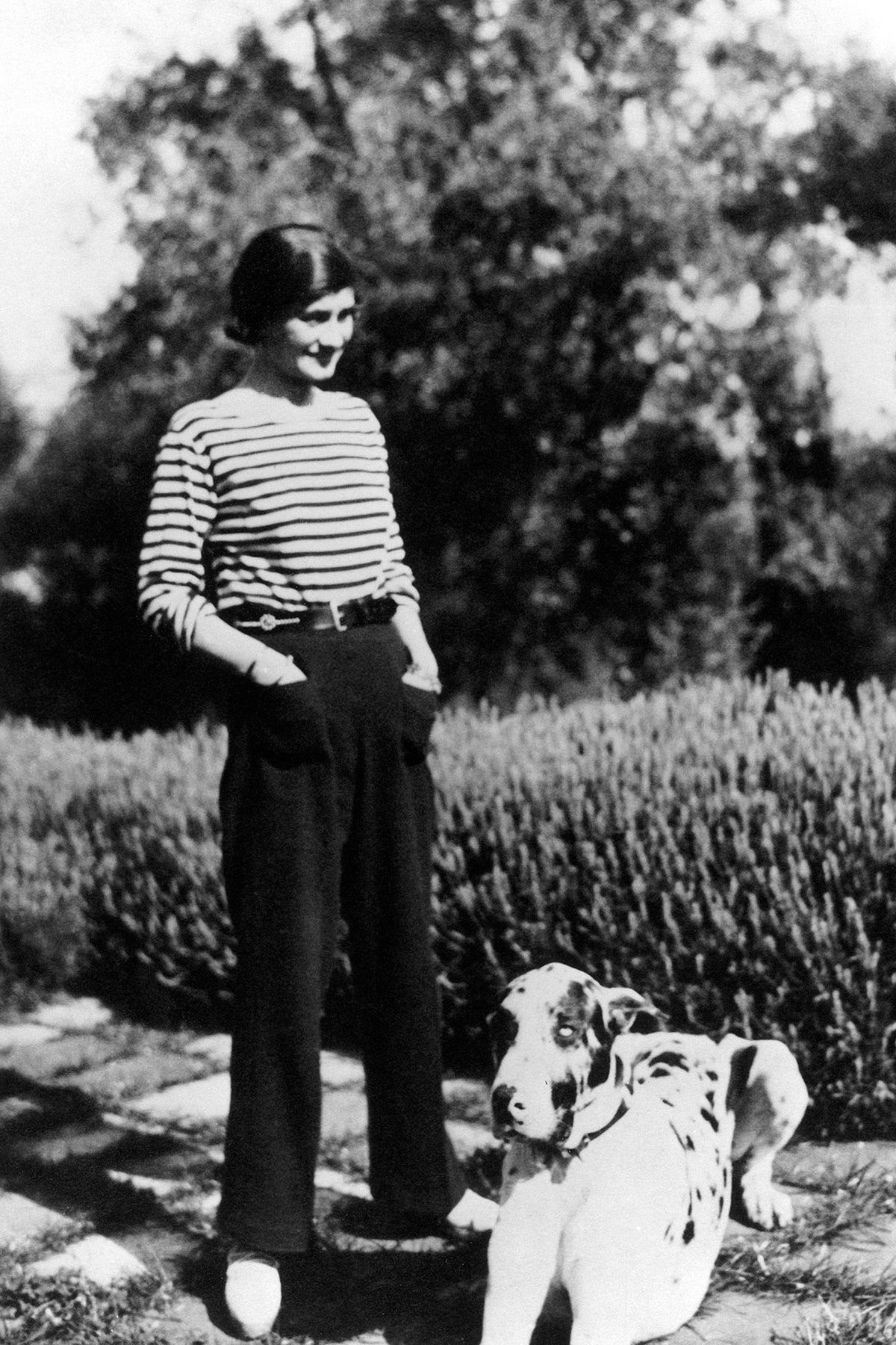 A woman in a striped shirt and dark trousers standing in a garden with a spotted dog.