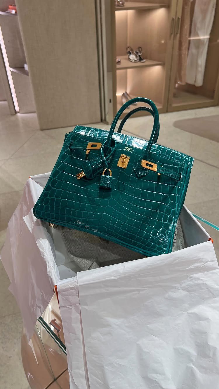 Teal crocodile leather handbag with gold hardware resting on white tissue paper inside a shopping bag.