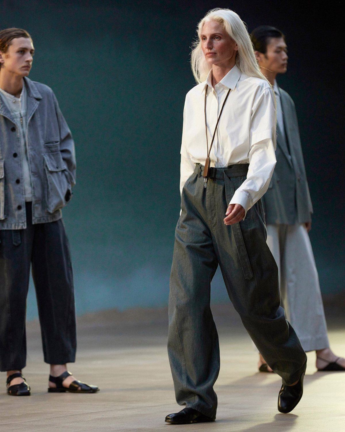 Models walking a runway wearing minimalist, high-fashion clothing in neutral tones against a dark teal background.