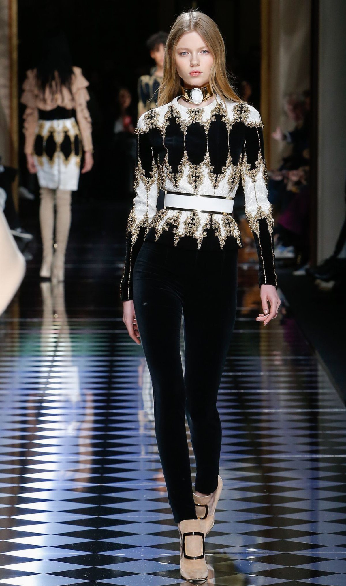 Blonde model walking a runway in an ornate black and gold top with black leggings.