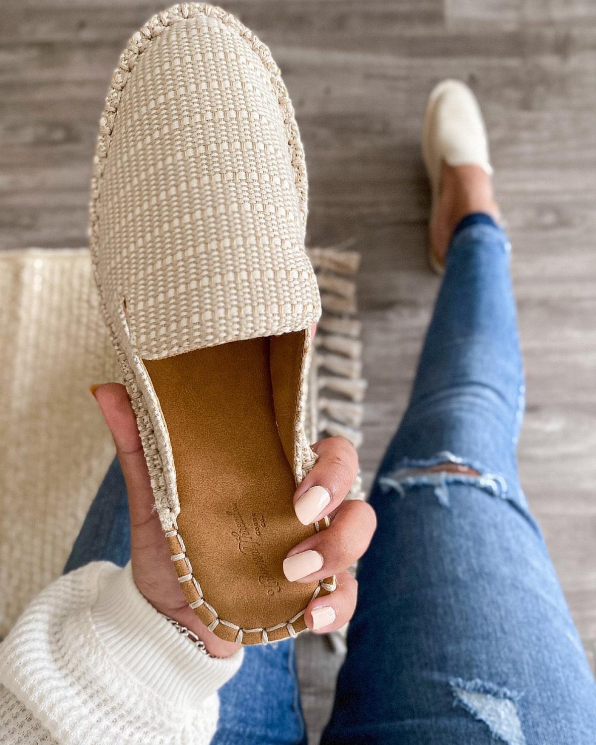 A person holding a beige woven espadrille shoe over their legs in blue jeans.
